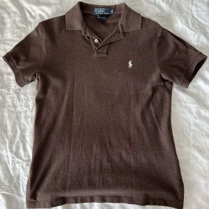 Ralph Lauren Polo Shirt - Säljer en klassisk brun pikétröja från Ralph Lauren med den ikoniska broderade logotypen på bröstet. Tröjan har korta ärmar och en stilren krage med knappar. Perfekt för en avslappnad men stilfull look.