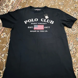 Svart t-shirt från Polo Club - Säljer en svart t-shirt från Polo Club med tryck på framsidan. Den har texten 'Polo Club Saint Vincent' och en amerikansk flagga. Perfekt för en casual look eller en avslappnad dag ute. T-shirten är i bomull och har korta ärmar. Ny med taggar!