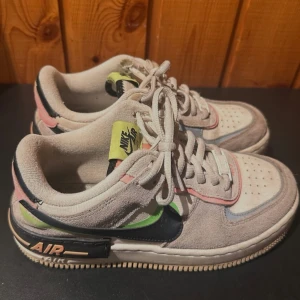 Nike Air Force 1 Shadow sneakers i pastellfärger - Säljer ett par Nike Air Force 1 Shadow sneakers i pastellfärger. De har en cool mix av beige, rosa och blå detaljer med klassisk snörning. Perfekta för vår och sommar! Skorna är i bra skick och redo för nya äventyr.