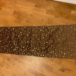 Brun bordsduk  med stjärnmönster - Säljer en superfin brun  bordsduk med glittrigt stjärnmönster. Den är 45x150 cm och gjord av 100% polyester. Perfekt för att liva upp en outfit med lite stjärnglans!✨