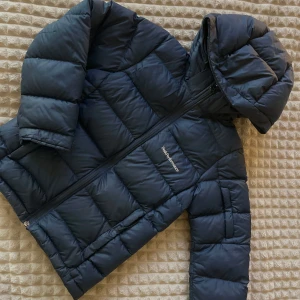 Svart dunjacka från Peak Performance JUNOR/BARN - Säljer en svart dunjacka från Peak Performance i JUNIOR STORLEK. Jackan är perfekt för vintern med sin pufferstil och långa ärmar. Den har en praktisk dragkedja och en mysig huva för extra värme. Perfekt för kalla dagar! 🖤 ”VARM, LÄTT OCH PACKBAR DUNJACKA.  ANVÄND SOM YTTERJACKA ELLER FÖRSTÄRKNINGSPLAGG I KALLA, TORRA FÖRHÅLLANDEN.” Avtagbar luva samt dragsko inuti längst ner. OBS! Finns ett litet hål på baksidan vid armbågen. Går säkert att fixa!   Använd men i fint skick. Nypris 2.700kr