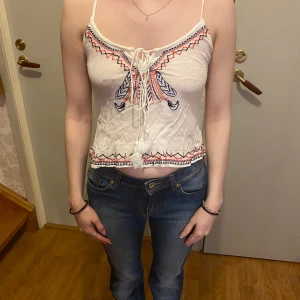 Vit broderad linnetopp - En fin vit linnetopp med broderade detaljer i rött och blått. Toppen har smala axelband och en söt knytning framtill med tofsar🤍