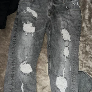 Grå slitna jeans - Säljer ett par grå jeans med coola slitningar och hål för en trendig look. De har en straight passform och är perfekta för en avslappnad stil. Jeansen har fem fickor och en klassisk dragkedja med knapp. Perfekta för höst och vår!
