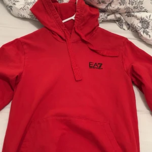 Röd hoodie från EA7 - Säljer en snygg röd hoodie från EA7. Den har en klassisk design med en känguruficka och justerbar huva. Perfekt för en avslappnad stil och passar bra till både vardag och träning. Märkesloggan är broderad på bröstet. Skön och bekväm att ha på sig! Passar storlek XS/S