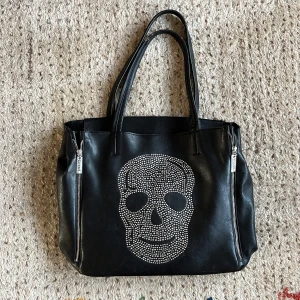 Svart väska med dödskallemotiv - Cool svart väska från Eternal med ett stort dödskallemotiv i silverfärgade nitar på framsidan. Väskan har två dragkedjor på sidorna och är gjord i syntetmaterial. Den har ett litet märke men är i bra skick.