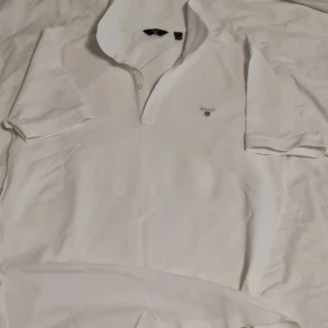 Vit kortärmad pikétröja från GANT - Säljer en klassisk vit pikétröja från GANT i storlek 16 år "Small". Tröjan är i bra skick och har en broderad logga på bröstet. Perfekt för både vardag och festliga tillfällen. Tillverkad i mjuk bomull för en bekväm passform.