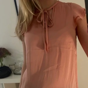 Rosa blus med knytband - Söt rosa blus i lätt och luftigt material. Den har korta ärmar och ett fint knytband vid halsen som ger en romantisk touch. Perfekt för både vardag och festliga tillfällen! Sparsamt använd 💗