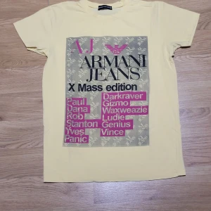 Gul T-shirt från Armani Jeans - Säljer en cool gul T-shirt från Armani Jeans med tryck i rosa och svart. Den har korta ärmar och ett stort grafiskt tryck på framsidan med texten 'X Mass edition'. Perfekt för en avslappnad stil eller fest! 🟡🖤