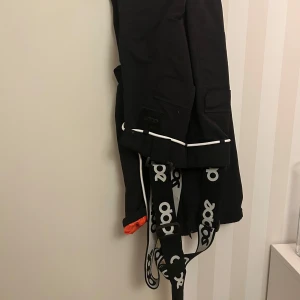 Svarta byxor med hängslen från Dope - Säljer ett par svarta byxor från Dope med coola hängslen och vita detaljer. Byxorna har praktiska fickor med knappar och en bekväm passform. Perfekta för vinterns äventyr! 🖤