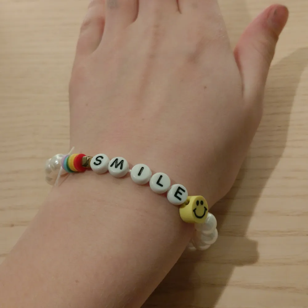 Superfint armband med vita pärlor och bokstavspärlor som bildar ordet 'SMILE'. Det har också en glad smiley och en regnbågsfärgad detalj. Perfekt för att sprida glädje och stil! 🥰. Accessoarer.