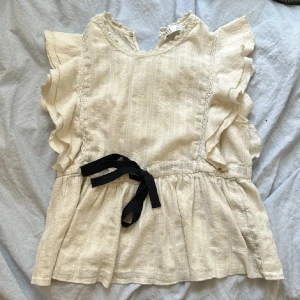 Beige blus med volanger från Anthropologie - Charmig beige blus från Anthropologie (Zara) i storlek S. Den har vackra volangdetaljer längs ärmarna och en söt knytning i midjan. Perfekt för vår och sommar, med en romantisk och luftig stil. Blusen är i mycket bra skick och redo för nya äventyr!