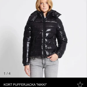 Jacka lager 157 - Snygg och glansig svart pufferjacka 'Nikki' i kort modell. Perfekt för kyliga dagar med sin varma vaddering och stilrena design. Jackan har en dragkedja framtill och långa ärmar. Passar perfekt till både vardag och fest!