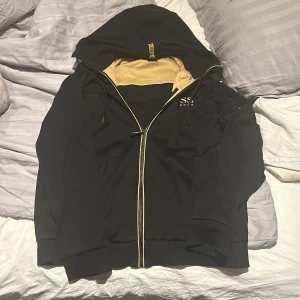 Svart zip hoodie från Hugo Boss - Säljer en snygg svart hoodie från Hugo Boss med gulddetaljer. Den har en dragkedja framtill och en stor huva. Perfekt för höst och vår, och den är i mycket bra skick. Passar både till vardags och lite finare tillfällen.