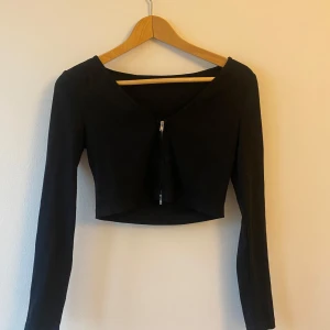 Svart croppad topp med dragkedja - Snygg svart croppad topp med långärmad design och dragkedja framtill. Toppen har en tight passform.