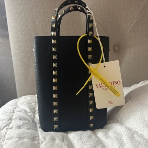 Svart handväska från Valentino Garavani  - Snygg svart handväska från Valentino i skinn med coola nitar längs handtagen. Perfekt för att ge din outfit en fin touch. Väskan är sprillans ny och har till och med lappen kvar, aldrig använd. Säljer för att inte riktigt passade min stil. En stilren design som passar både till vardags och fest. Pris kan diskuteras.