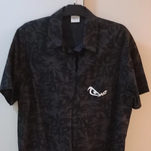 Svart kortärmad skjorta med mönster - Säljer en svart kortärmad skjorta med ett subtilt mönster och en vit broderad detalj på bröstet. Perfekt för en avslappnad sommarlook eller en utekväll. Skjortan har en klassisk krage och knappar framtill. Passar bra till både jeans och shorts!