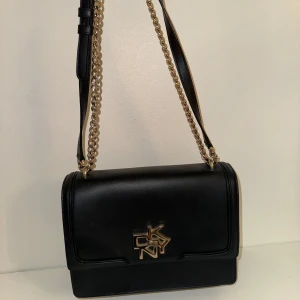 Svart axelväska från DKNY - Snygg svart axelväska från DKNY i skinn med guldfärgade kedjedetaljer. Väskan har en stilren design med ett stort DKNY-emblem på framsidan. Insidan är fodrad med logotypmönster och har en praktisk dragkedjeficka. Perfekt för både vardag och fest!