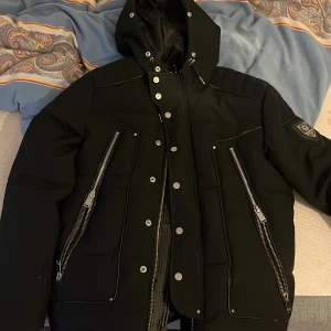 Svart dunjacka från Horspist - Säljer en snygg svart dunjacka från Horspist, perfekt för vintern. Jackan har en cool design med dragkedjor och knappar framtill. Den är långärmad och har en huva för extra värme. Märkesdetaljer på ärmen ger en lyxig touch. Passar både till vardags och fest!