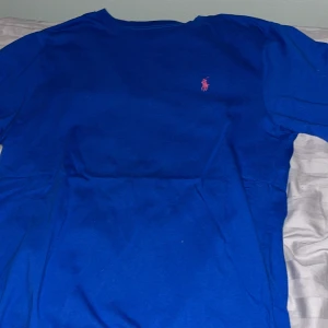 Blå t-shirt från Polo Ralph Lauren - Säljer en snygg blå t-shirt från Polo Ralph Lauren. Den har en klassisk design med korta ärmar och en liten rosa broderad logga på bröstet. Perfekt för en avslappnad stil eller sommarens äventyr. Passar bra till jeans eller shorts!