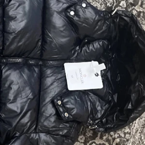 Svart dunjacka från Moncler - Säljer en snygg svart dunjacka från Moncler i storlek M. Jackan är i mycket bra skick och perfekt för vintern. Den har en dragkedja framtill och är långärmad med en huva för extra värme. Passar perfekt för kalla dagar!