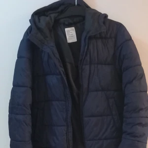 Mörkblå quiltad jacka - Säljer en mörkblå quiltad jacka i mycket bra skick. Den är perfekt för höst och vinter med sin varma vaddering. Jackan har en dragkedja framtill och en bekväm huva. Passar både till vardags och lite mer avslappnade tillfällen. Storleken är inte angiven men ser ut att vara normal passform.
