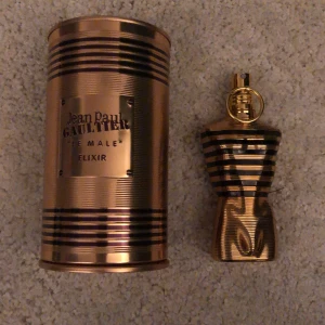 Jean Paul Gaultier Le Male Elixir Parfym - Säljer en elegant parfymflaska från Jean Paul Gaultier, Le Male Elixir. Flaskan har en unik design som liknar en manlig torso med guld- och svartfärgade ränder. Den kommer i en snygg metallcylinder med samma färgtema. Perfekt för den som vill ha en lyxig och maskulin doft. Ungefär 65ml kvar.