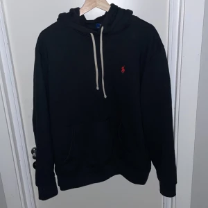 Svart hoodie från Ralph Lauren - Säljer en klassisk svart hoodie från Ralph Lauren med det ikoniska röda logotypen på bröstet. Den har en justerbar huva med vita snören och en praktisk magficka. Perfekt för både vardag och chill. Passar till allt och är superbekväm! 🖤