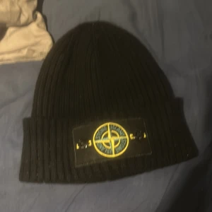 Svart ribbad mössa från Stone Island - Säljer en snygg svart ribbad mössa från Stone Island. Den är tillverkad i Italien och har en cool broderad logga framtill. Perfekt för att hålla sig varm och stilren under kyliga dagar. Passar både till vardags och mer uppklädda tillfällen.