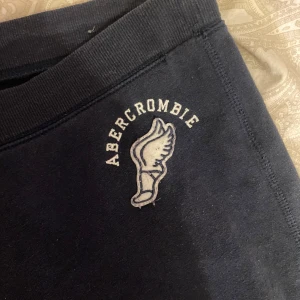 Abercrombie & Fitch - Säljer ett par mörkblå mjukisbyxor från Abercrombie & Fitch med snörning i midjan och ett broderat märke på sidan. 