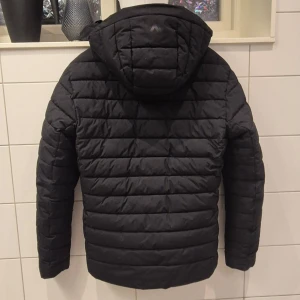 Svart dunjacka från McKinley - Säljer en svart dunjacka från McKinley med huva och dragkedja. Jackan har en quiltad design och långa ärmar, perfekt för kyligare väder. Den är i storlek small och passar bra för både höst och vinter.
