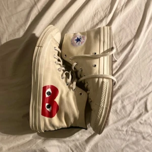 Converse x Comme des Garçons - Snygga beige höga sneakers med det ikoniska röda hjärtat från Comme des Garçons. Skorna har klassisk snörning och Converse All Star-logga på sidan. Perfekta för en stilren look.