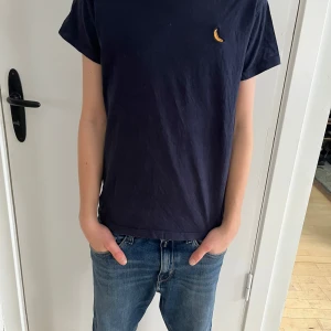 Mörkblå t-shirt med bananbroderi - Säljer en marinblå T-shirt i storlek 150 cm (passar barn 9–12 år). T-shirten har en rund halsringning och ett litet broderat bananmotiv på bröstet. Materialet är mjukt och skönt, perfekt för vardagsbruk.  Personen på bilderna är 160 cm lång, 12 år gammal, och har en smal/normal kroppsbyggnad.  Skick: Bra begagnat skick, med minimala tecken på användning.  Nypris: 200 SEK