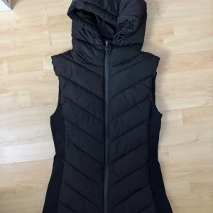 Svart dunväst från Zara - Snygg svart dunväst från Zara med huva och dragkedja. Perfekt för kyliga dagar och ger en stilren look. Västen har en quiltad design som ger extra värme och komfort.