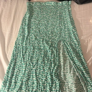 Grön mönstrad långkjol - Säljer en grön långkjol med ett vitt mönster. Kjolen har en slits som ger en fin detalj och rörelse. Perfekt för varmare dagar och ger en luftig känsla.