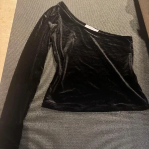 Svart oneshoulder topp från Costes Studio - Snygg svart oneshoulder topp från Costes Studio i sammet. Perfekt för en kväll ute med sin eleganta design i velour. Toppen har en lång ärm och en stilren look.