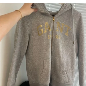 Grå hoodie från GANT - Snygg grå hoodie från GANT med dragkedja och guldtryck på bröstet. Den har en justerbar huva med snören och praktiska fickor framtill. Perfekt för en avslappnad stil.