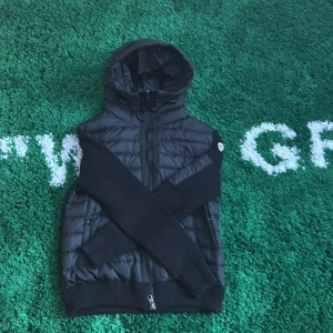 Svart quiltad jacka från Moncler - Snygg svart quiltad jacka från Moncler med huva och dragkedja. Jackan har långa ärmar i stickat material och en broderad logotyp på ärmen. Perfekt för kyligare dagar.