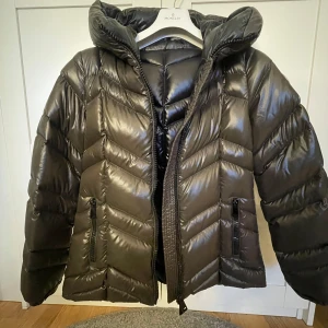 Pufferjacka från Moncler - Snygg svart pufferjacka från Moncler med glansig finish och huva. Jackan har en dragkedja framtill och Moncler-logga på ärmen. Perfekt för kyliga dagar. Storlek 3