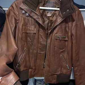 Brun skinnjacka från JSFN/ellos - Snygg brun skinnjacka från JSFN/ellos med dragkedja och knappar. Jackan har flera praktiska fickor med dragkedjor och en hög krage med knäppning. Perfekt för en cool och stilren look.