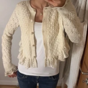 Beige stickad kofta - Säljer en söt beige stickad kofta med knappar framtill. Koftan har ett fint mönster och volangdetaljer vid nederkanten och på baksidan.