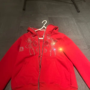 Röd hoodie med glitterdetaljer från Le Dos - Snygg röd hoodie från Le Dos med glittriga motiv och text på framsidan. Tröjan har dragkedja och en mysig huva. Perfekt för att sticka ut med lite extra bling! 🔥