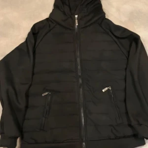 Svart pufferjacka med huva - Snygg svart pufferjacka med huva och dragkedja framtill. Jackan har långa ärmar och två praktiska fickor med dragkedjor. Perfekt för kyligare dagar.Aldrig använd och nypris 1000:-