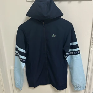 Blå och ljusblå jacka från Lacoste - Snygg blå och ljusblå jacka från Lacoste med dragkedja och huva. Jackan har långa ärmar med Lacoste-logga på ärmarna och bröstet. Perfekt för en sportig look. Skick 8/10, Org pris 2000 mitt pris 1000 priset går att diskuteras, storlek xs-S 