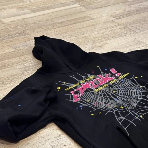 Svart hoodie med spindelnätsdesign - Cool svart hoodie med en stor spindelnätsdesign och text i rosa och gult på framsidan. Baksidan har en liten gul stjärna. Perfekt för en avslappnad stil med en touch av edge. Endast: 299