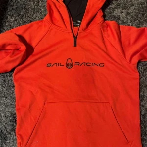 Röd hoodie från Sail Racing - Säljer en snygg röd hoodie från Sail Racing limited edition med svart logga på bröstet. Den har en praktisk dragkedja vid halsen och en stor ficka framtill. Perfekt för kyliga dagar. Köpt för 2200