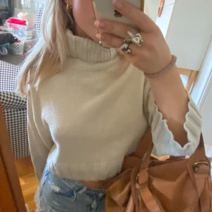 Beige croppad stickad polotröja - Säljer en snygg beige croppad stickad tröja med hög krage. Perfekt för en trendig look med långa ärmar och ribbade detaljer vid ärmslut. Passar utmärkt till jeans eller kjol för en avslappnad stil.