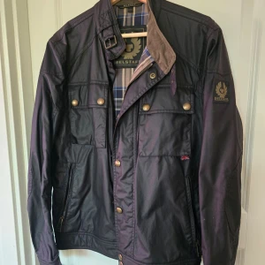 Svart jacka från Belstaff - Säljer en stilren svart jacka från Belstaff med knappar och dragkedja. Jackan har en klassisk design med flera fickor och ett rutigt foder. Perfekt för höst och vår.