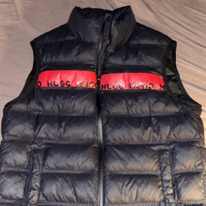 Svart dunväst från Hugo Boss - Snygg svart dunväst från Hugo Boss med röd detalj och logotyp på bröstet och ryggen. Västen har en dragkedja framtill och är perfekt för kyligare dagar.