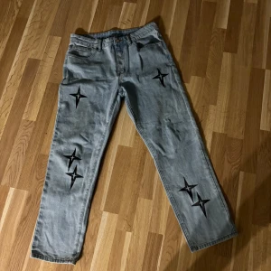 Grå jeans med stjärndetaljer - Snygga grå jeans med unika svarta stjärndetaljer på benen. Perfekt för dig som vill sticka ut med en cool och trendig stil. De har en klassisk femficksdesign och en bekväm passform.