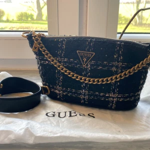 Axelväska från Guess - Rutig axelväska från Guess med guldfärgade kedjedetaljer och dragkedja. Väskan är i ett tjockare tyg med mönster och två stycken medföljande axelremmar. Insidan är fodrad med ett mönstrat tyg och har flera fack för enkel organisering. Väskan är i nyskick, aldrig använd och därmed fläckfri. Dustbag och extra läder rem medföljer. Inköpt i Guess butik.